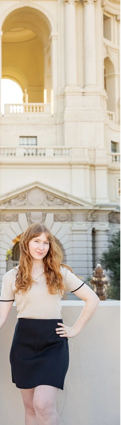 A woman with red hair standing outdoors in front of a large, elegant building with classical architecture, including arches and columns, during daylight.