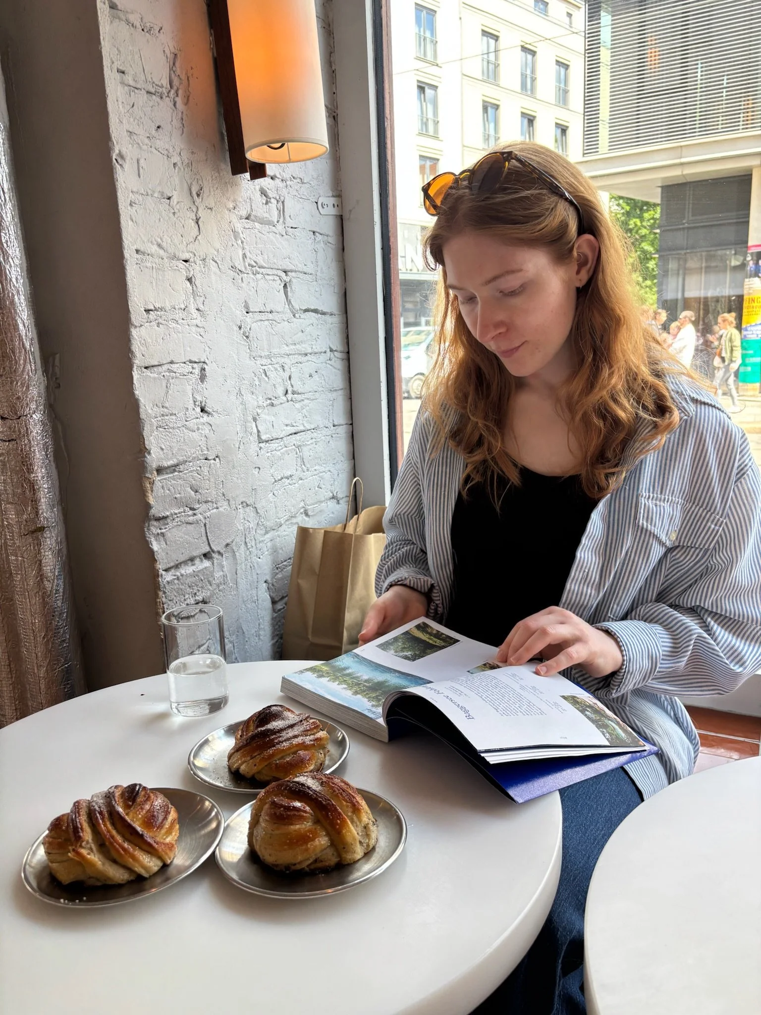 A young woman with red hair and sunglasses on her head sitting at a table by a large window in a cafe. She is looking at a book. On the table, there are three trays with pastries, a glass of water, and a blue notebook.