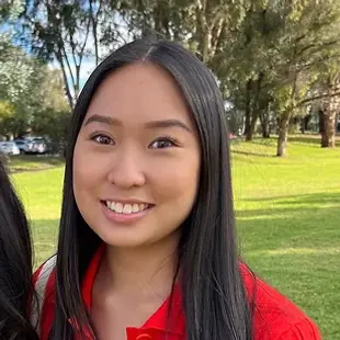Smiling woman with long hair wearing a red shirt, standing outdoors in a grassy area with trees.