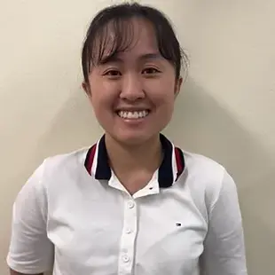 Smiling person wearing a white shirt with a dark collar standing against a light-colored wall.