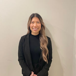 A person wearing a black blazer and long hair, smiling in front of a plain wall.