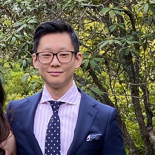 Man in suit and tie wearing glasses in an outdoor setting with greenery in the background.