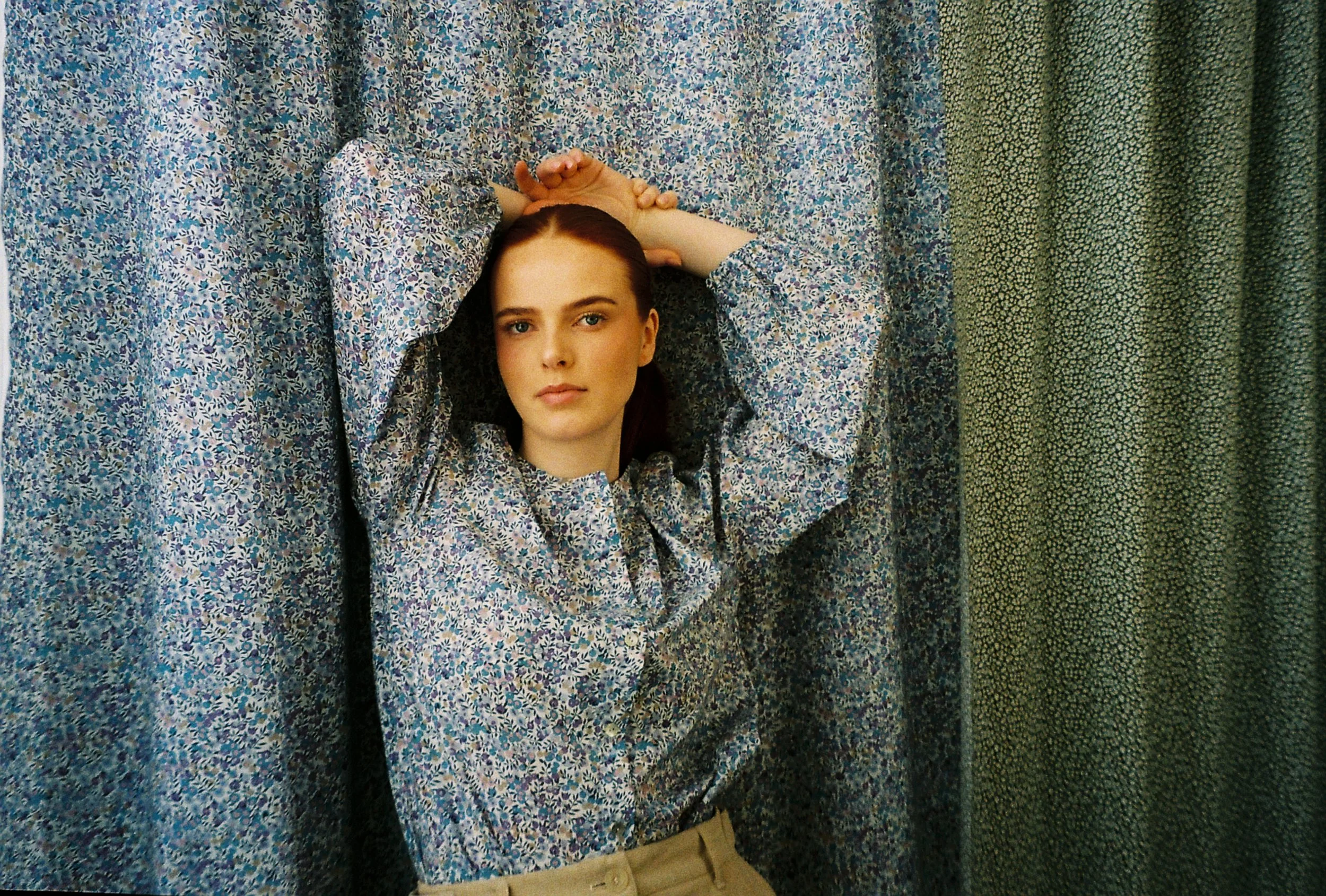 En ung kvinne med brunt hår og lys hud står foran gardiner med blå og grønne mønstre, med armene løftet over hodet, iført en blå blomstret skjorte.