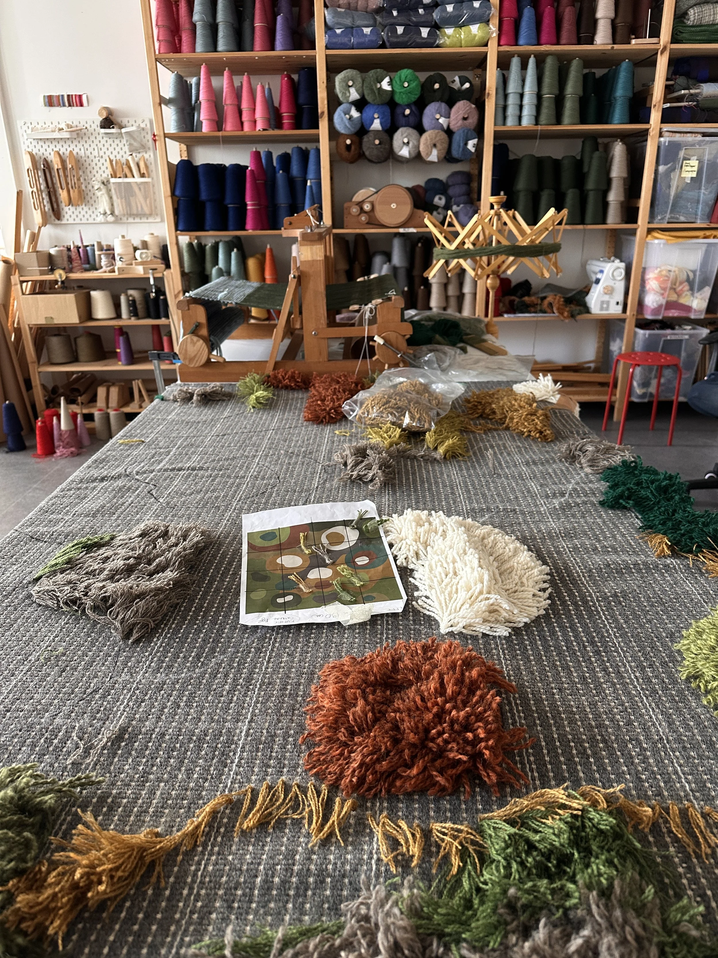 Arbeidsbord med teppeknyting, omgitt av hyller med garnnøster i ulike farger.