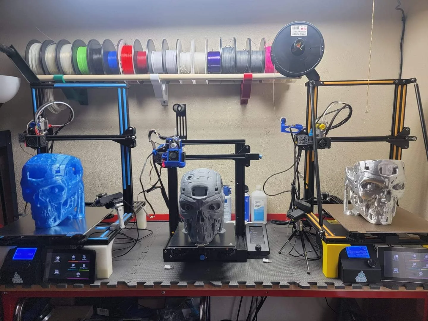 Three 3D printers on a workbench, each printing a detailed skull model, with filament spools on a shelf above.