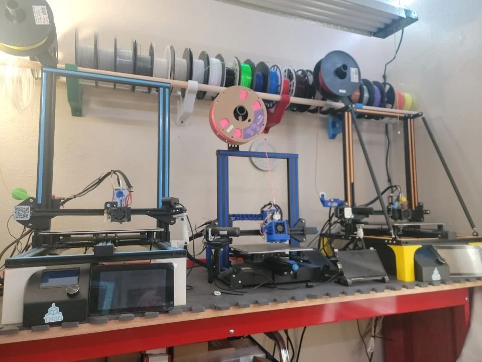 Three 3D printers on a workbench with various spools of filament on a shelf above.
