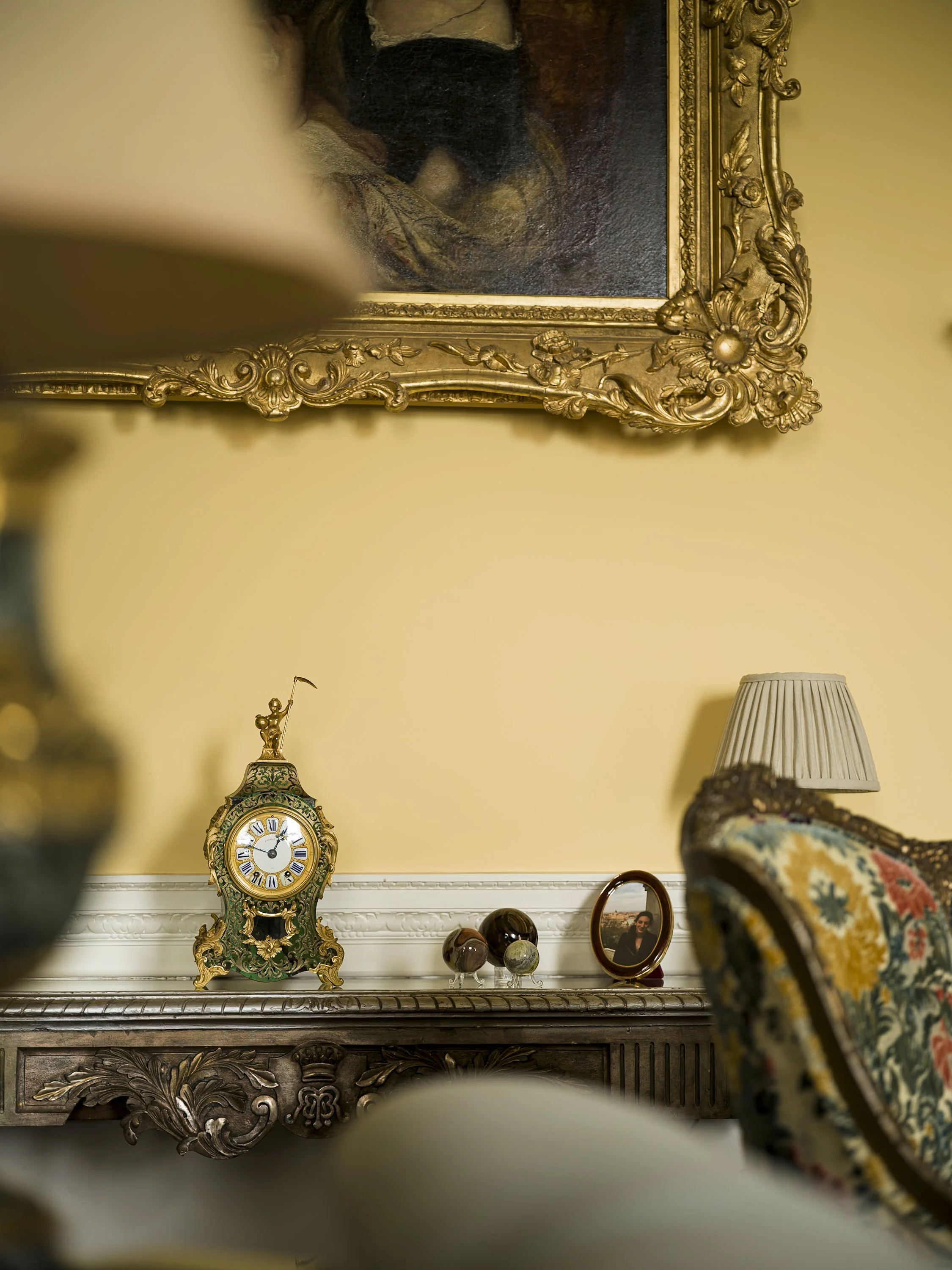 A vintage clock, a framed photograph, and decorative glass spheres on a dark ornate wooden table against a yellow wall with a large ornate gold-framed painting hanging above.