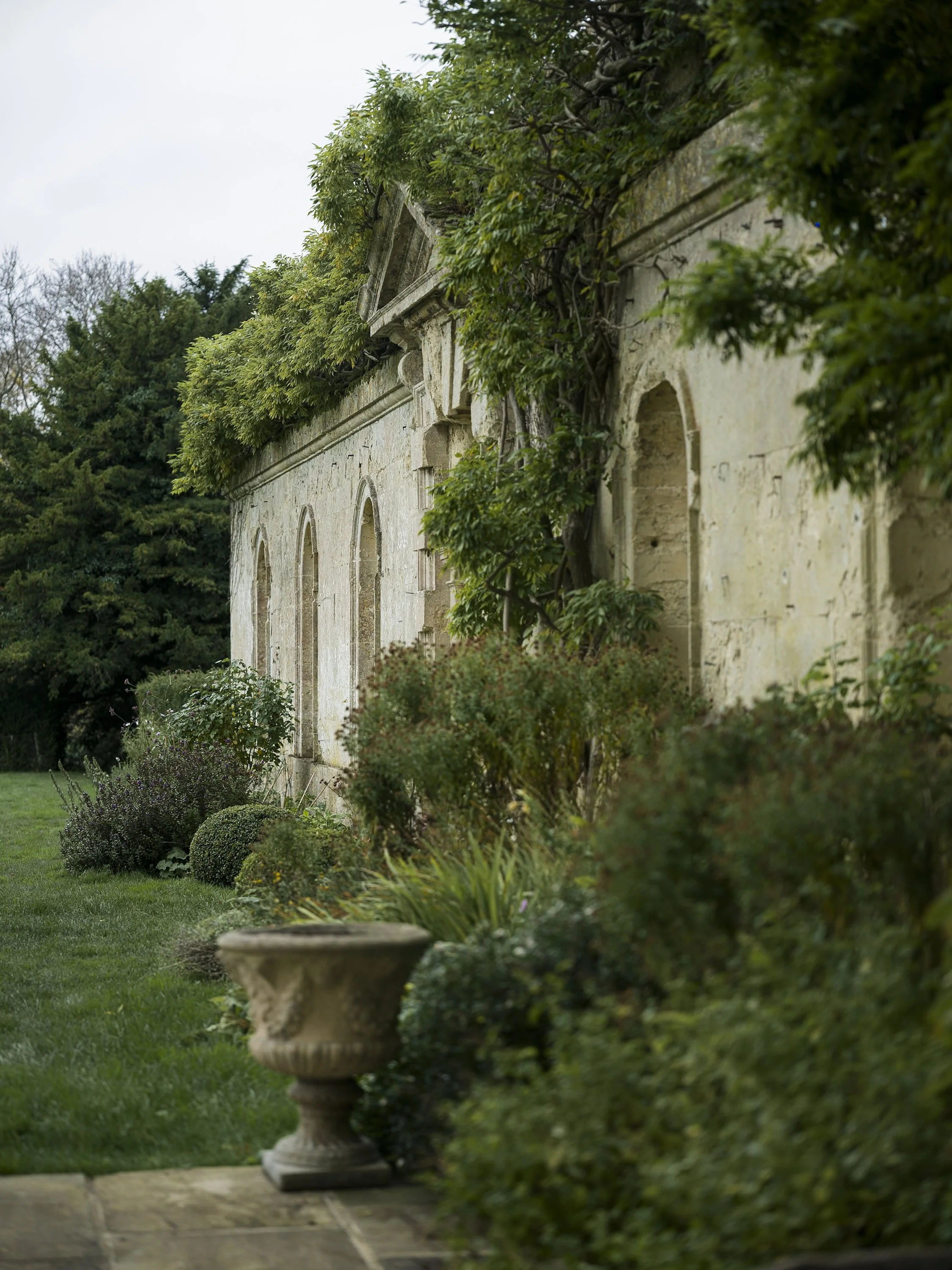 An old stone building with arched windows, surrounded by lush green plants and shrubs, with a stone urn in the foreground.