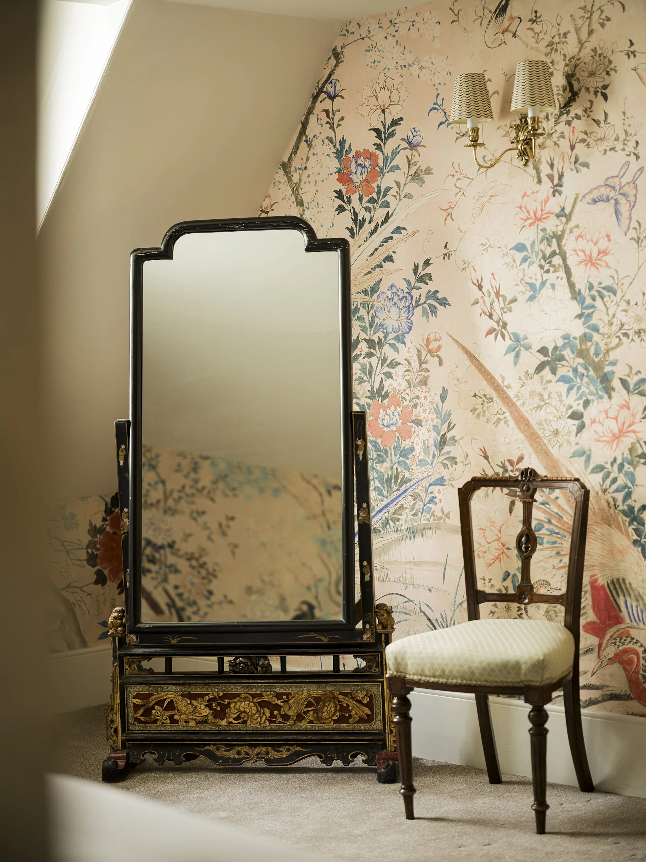 Room with floral wallpaper, a black and gold antique dressing mirror, an ornate chair with a cream upholstered seat, and a wall-mounted gold and cream lampshade fixture.