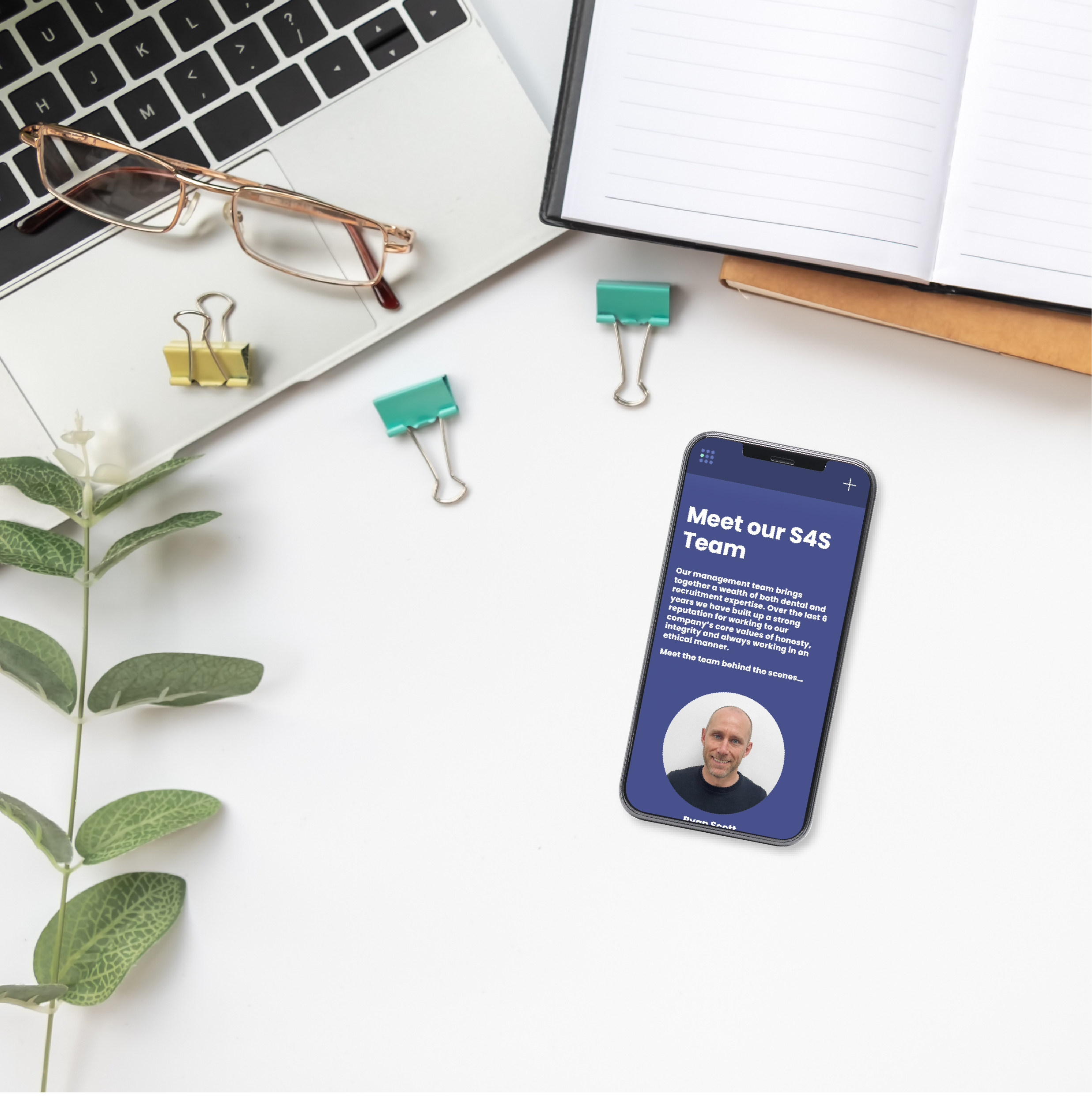 Laptop, glasses, binder clips, plant, open notebook, smartphone displaying "Meet our S4S Team" on a white surface.