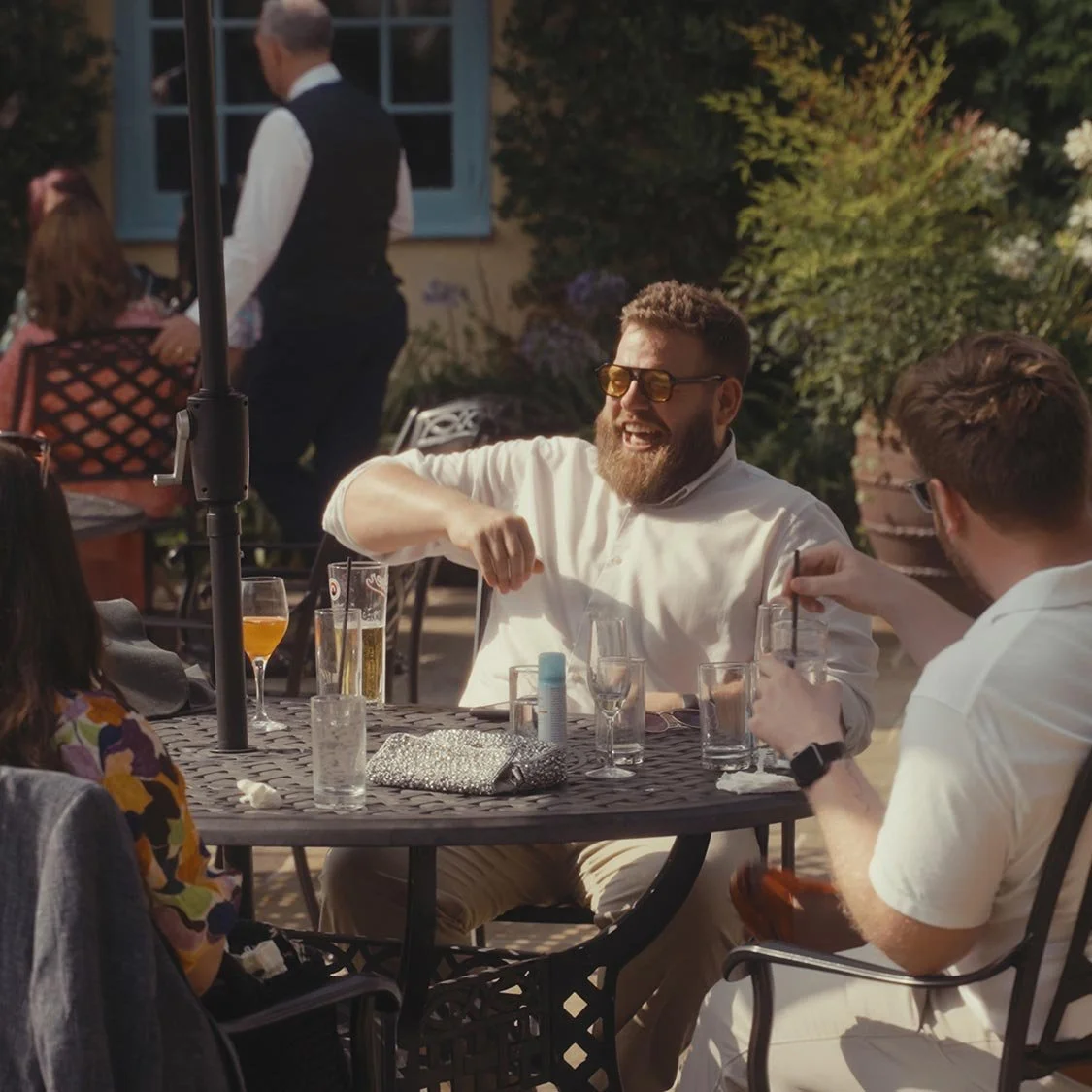 Group of people sitting at an outdoor table, socializing, with drinks and glasses on the table, garden setting with plants and flowers in the background.