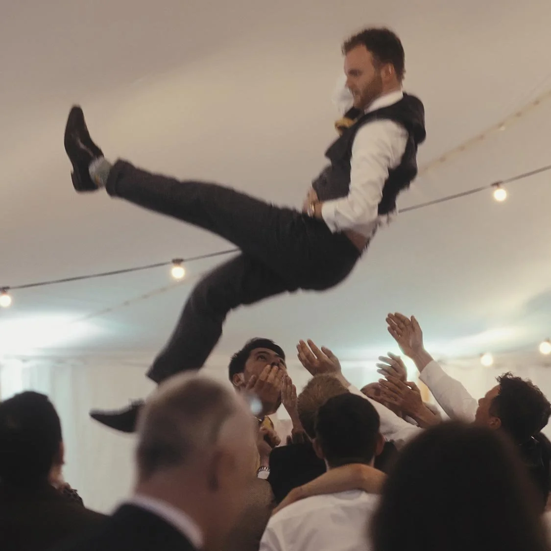 A man being thrown into the air by a crowd at a celebration event under string lights in a tent.