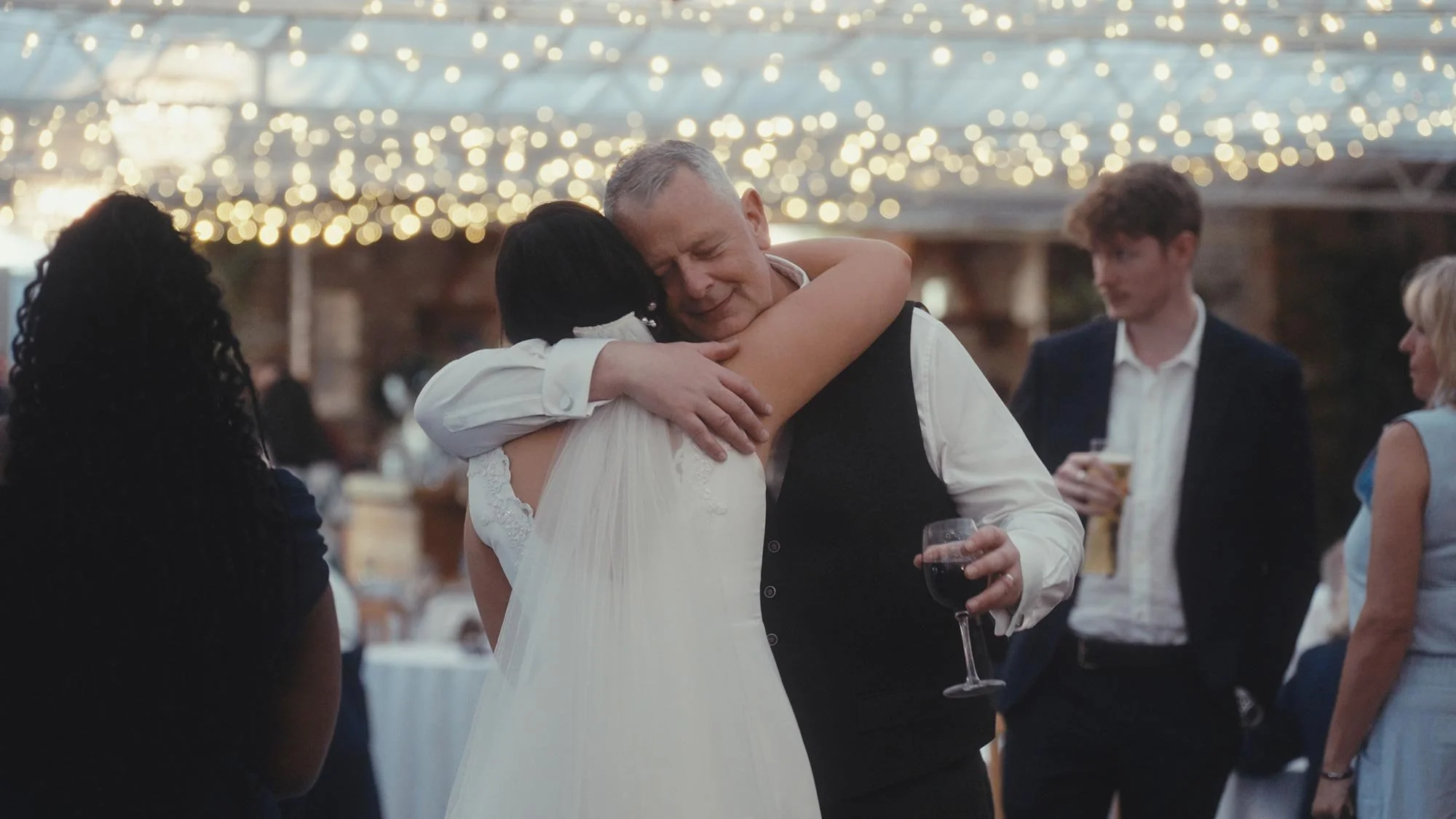 A daughter and her father hugging at a wedding, with people standing nearby holding drinks, under a ceiling decorated with string lights.