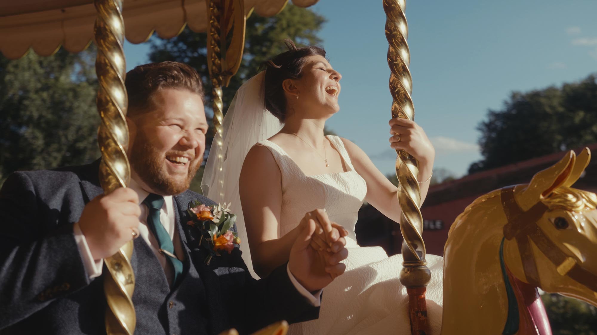 A smiling bride and groom riding a carousel horse outdoors on a bright, sunny day. The groom is holding the pole of the carousel, dressed in a dark suit with a boutonnière, and the bride is wearing a white wedding dress and a veil, holding onto the pole with a joyful expression.