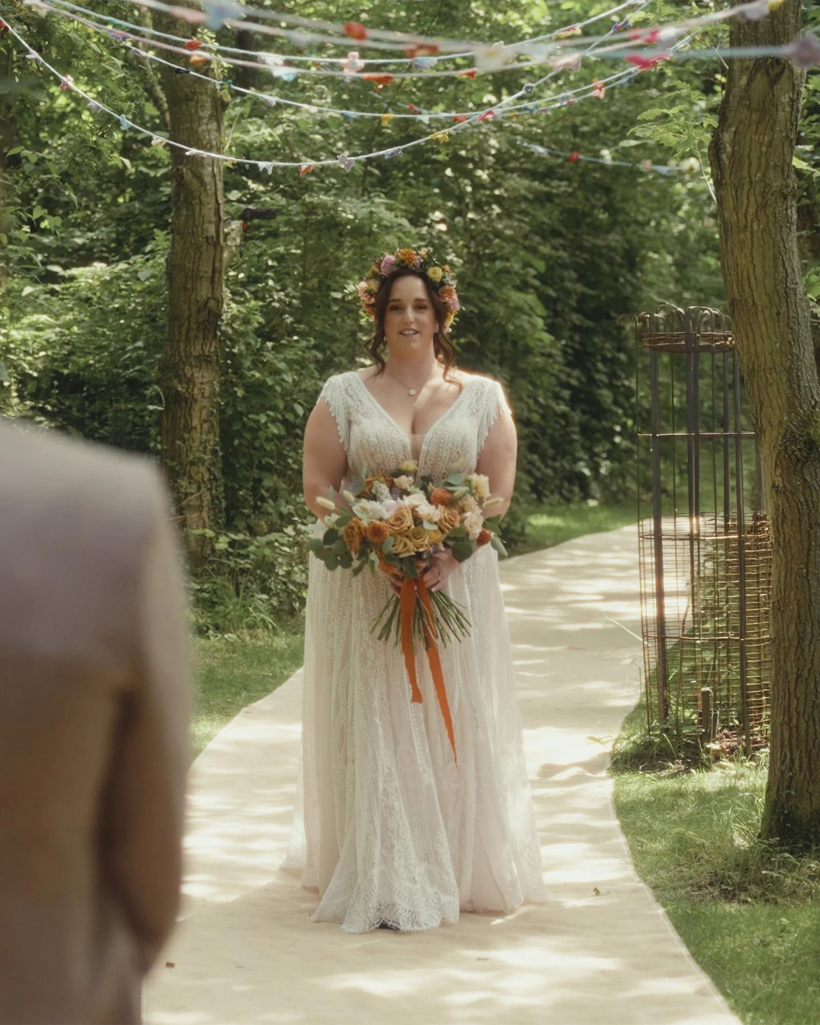 A bride in a white wedding dress holding a bouquet of flowers stands outdoors on a path surrounded by trees. Floral decorations hang overhead, and her groom stands facing her in the foreground.