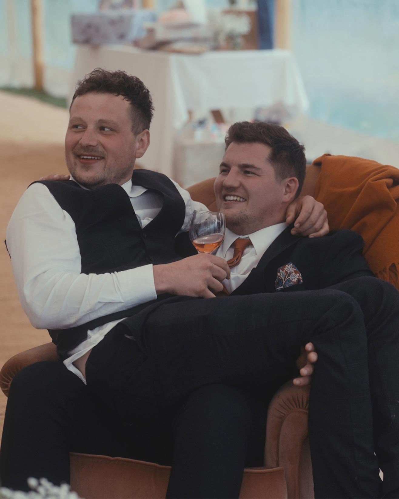 Two men sitting on a chair in a relaxed setting, one holding a glass of wine.