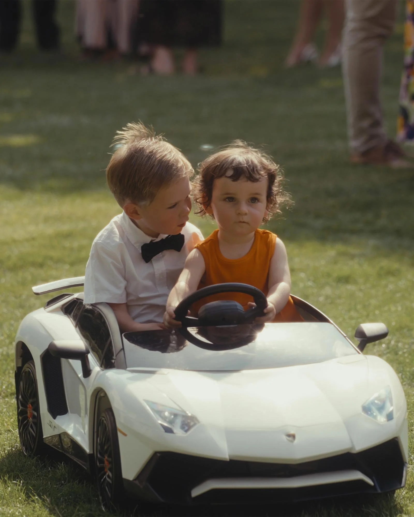 Two young children riding in a toy white sports car on a grassy lawn, with people blurred in the background.