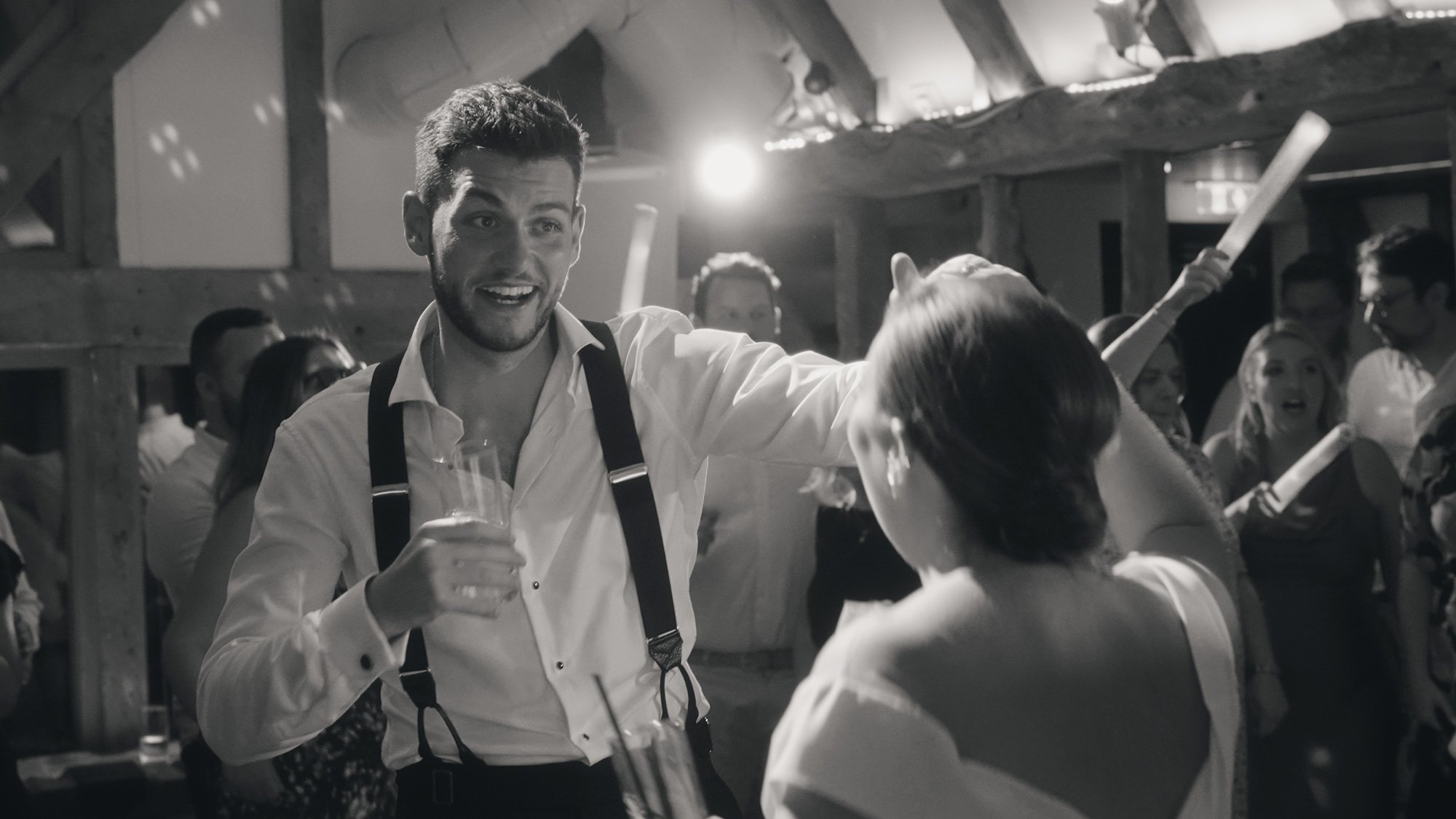 Black and white photo of a lively party scene with people dancing, a man in a shirt with suspenders smiling while holding a drink.