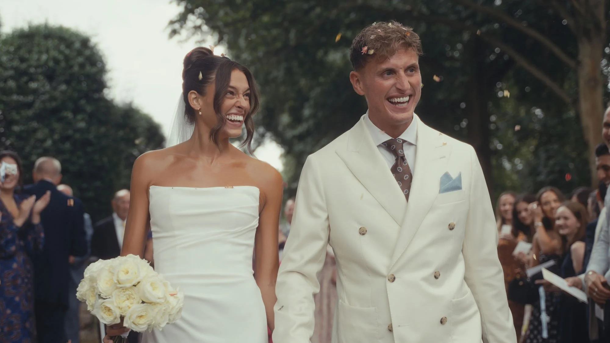 A bride and groom are smiling and holding hands at their wedding ceremony outdoors, surrounded by friends and family.