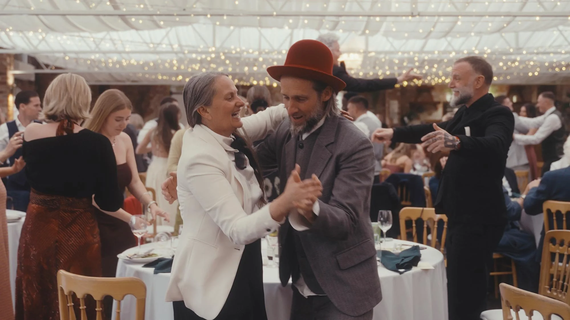 People dancing and socializing at a wedding reception under hanging string lights.
