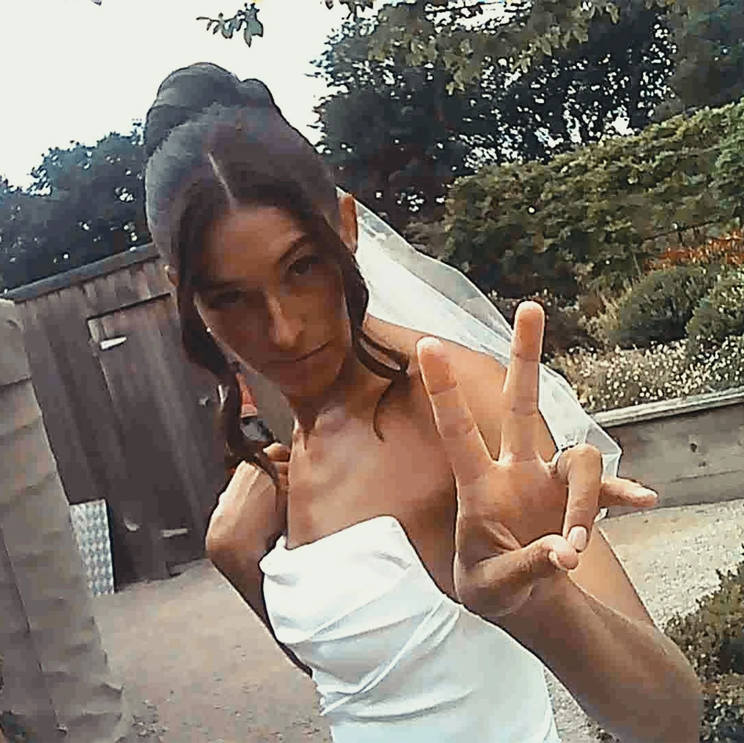 A woman with dark hair styled in an updo, wearing a strapless white dress with a veil, making a peace sign with her right hand.