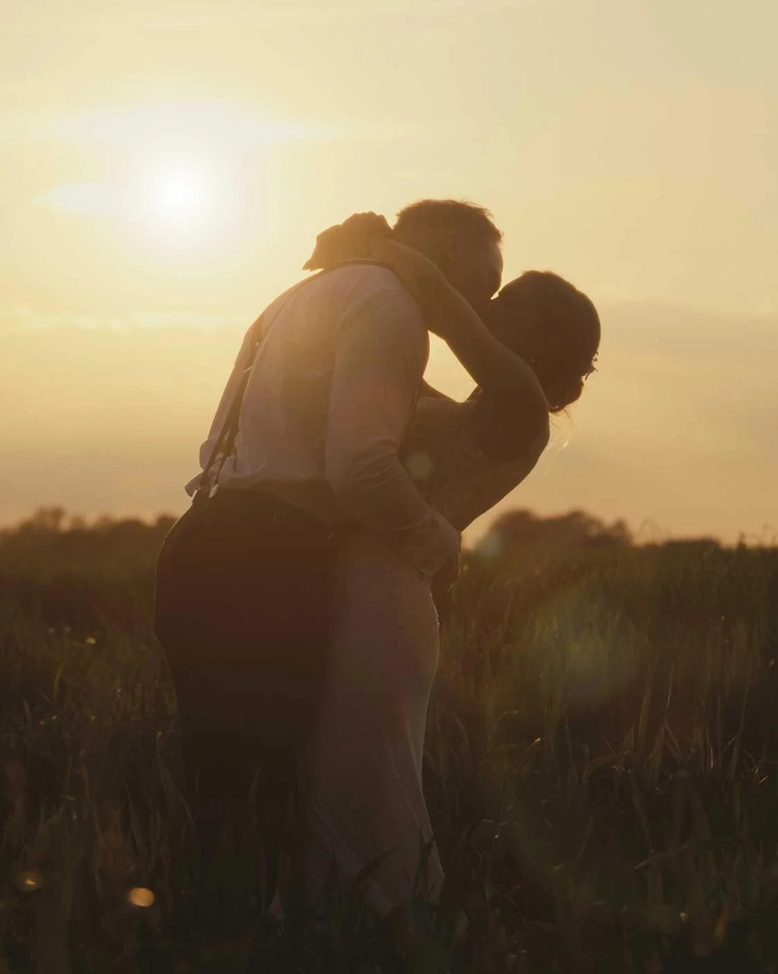 Silhouetted couple embracing and kissing in a field at sunset.
