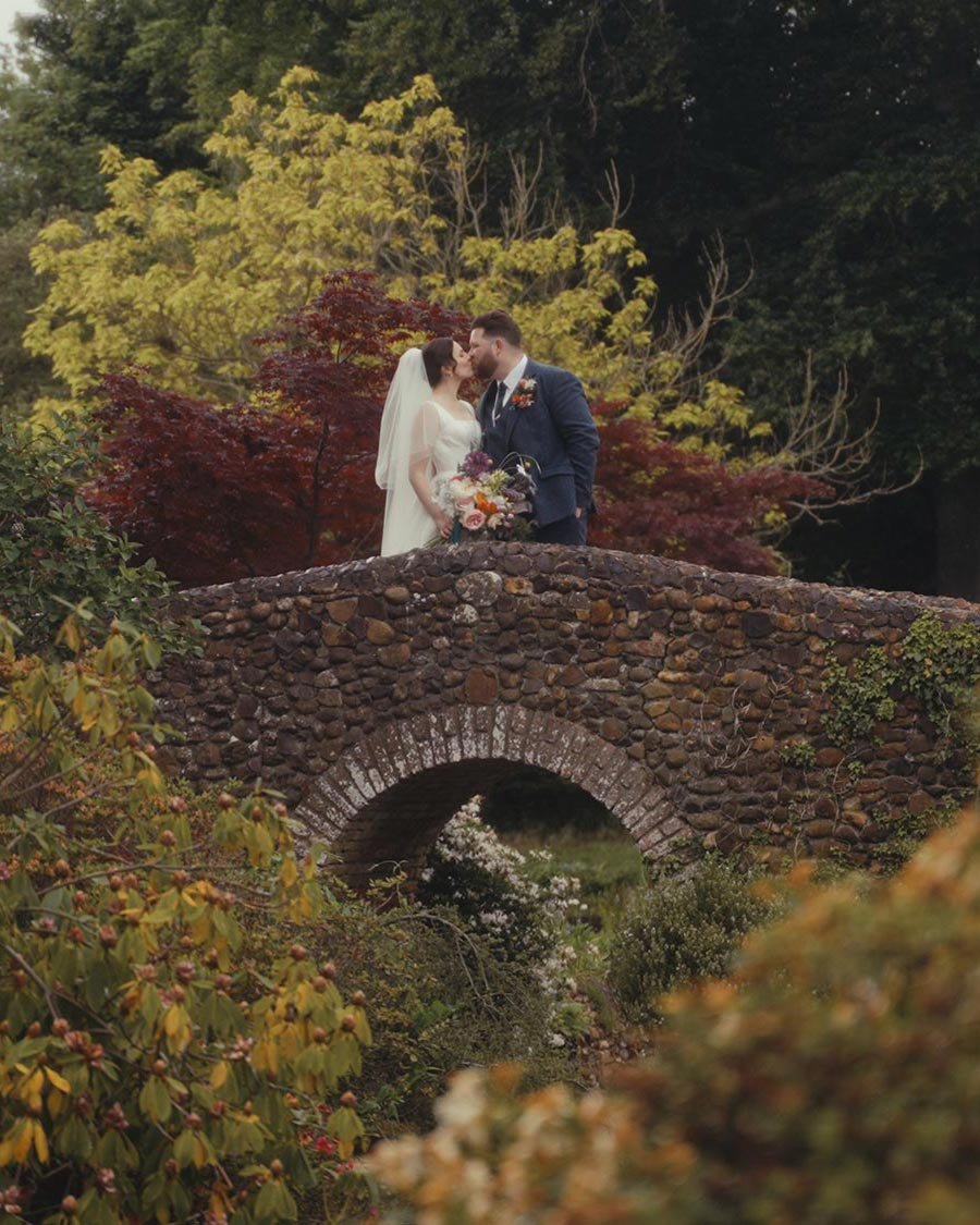 A bride and groom share a kiss on a stone bridge surrounded by lush, colorful garden foliage.