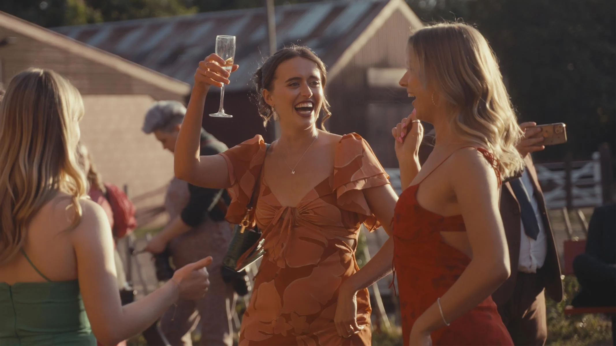 Three women at an outdoor gathering, one with a champagne glass and a joyful expression, the others conversing and dressed in elegant dresses, with natural sunlight and rustic buildings in the background.