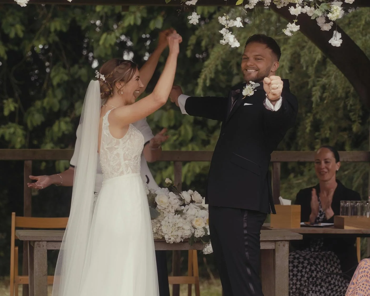 Bride and groom celebrating at an outdoor wedding ceremony, surrounded by flower decorations and applauding guests.