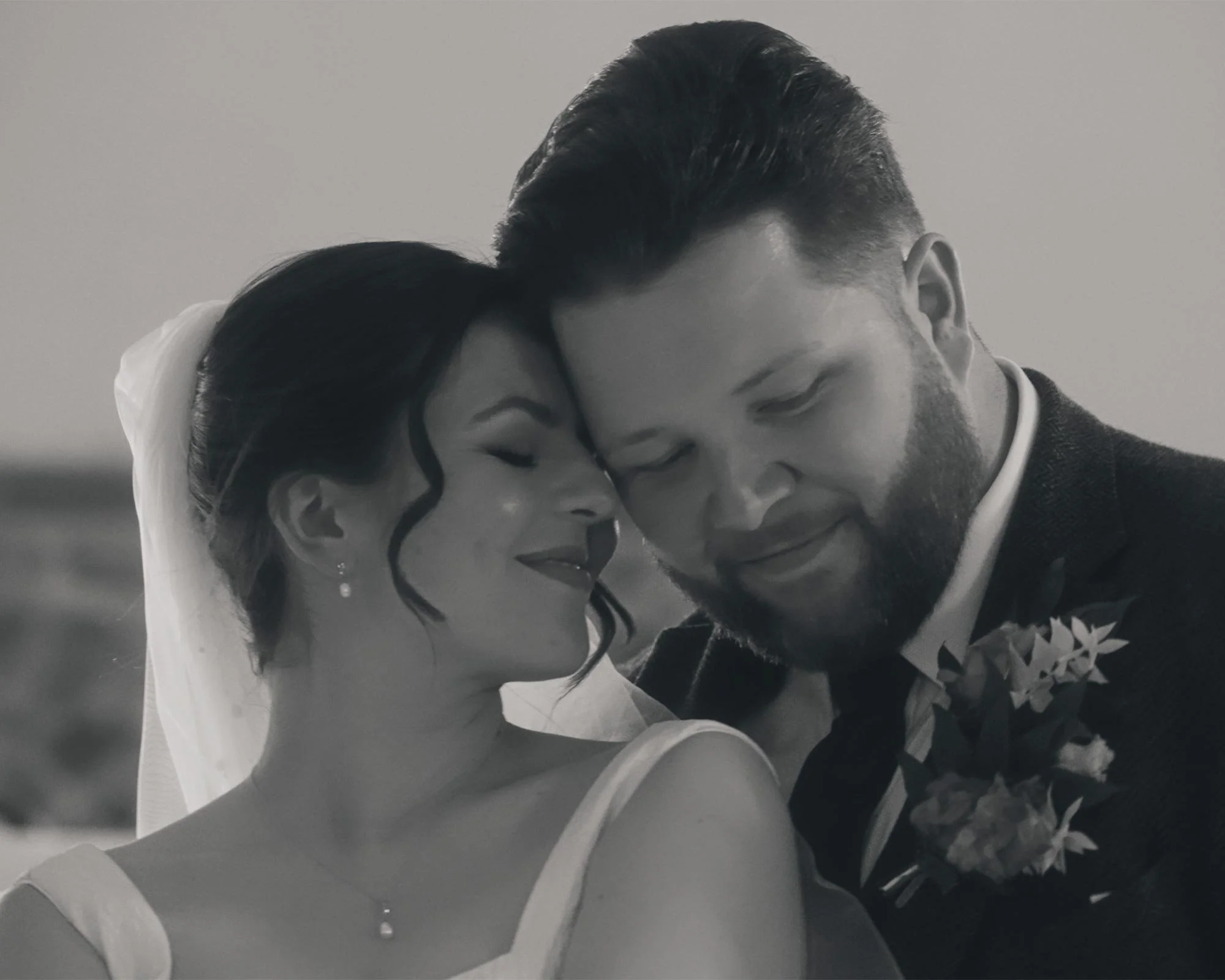 A bride and groom in wedding attire sharing a tender moment, their foreheads touching, eyes closed, smiling gently.