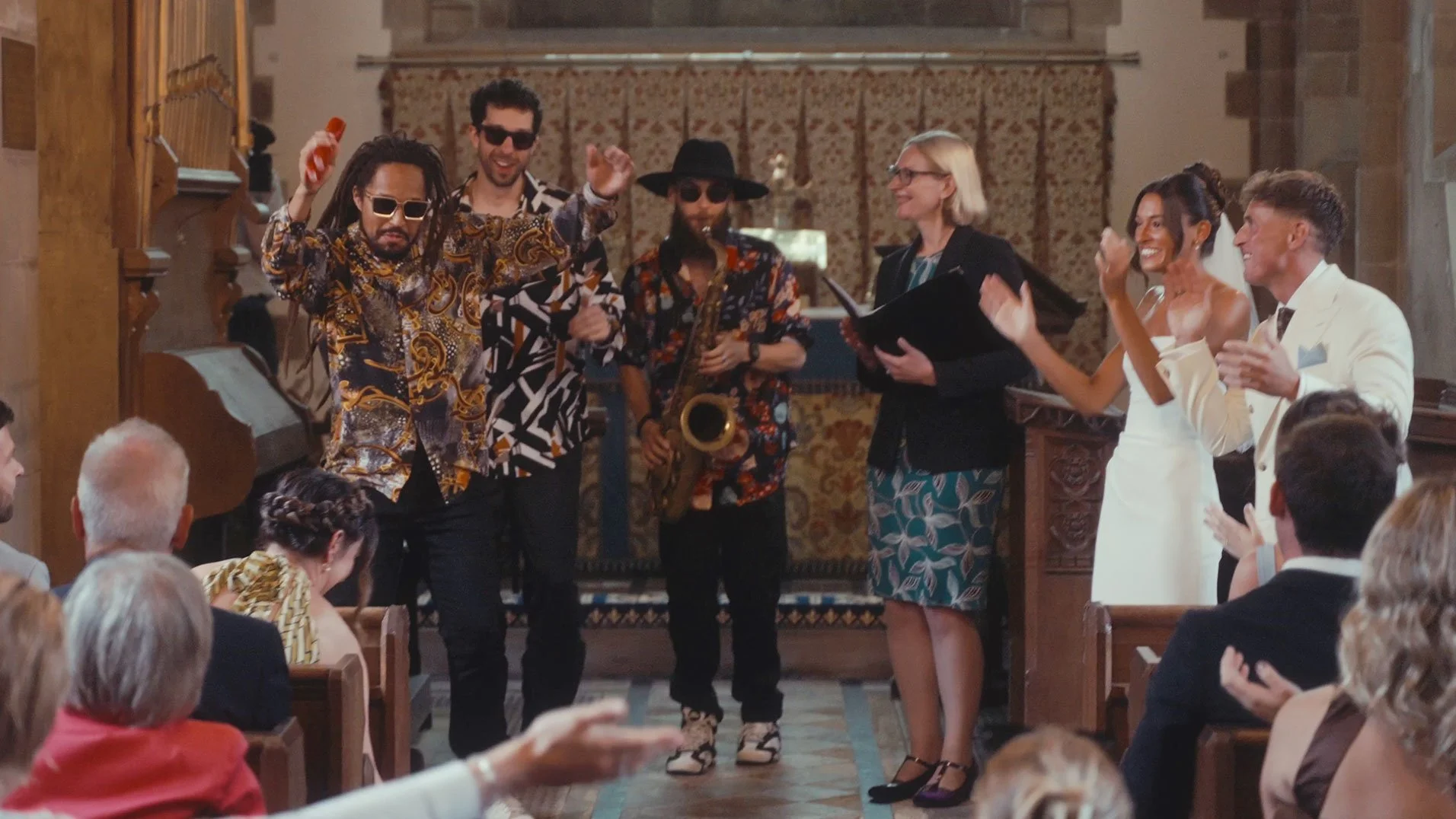 A group of musicians and a singer performing at a wedding ceremony in a church. The band includes a man with dreadlocks wearing sunglasses, a man with a saxophone, and another man with a hat and sunglasses. The bride and groom are clapping and smiling in front of the band, with wedding guests seated and applauding.
