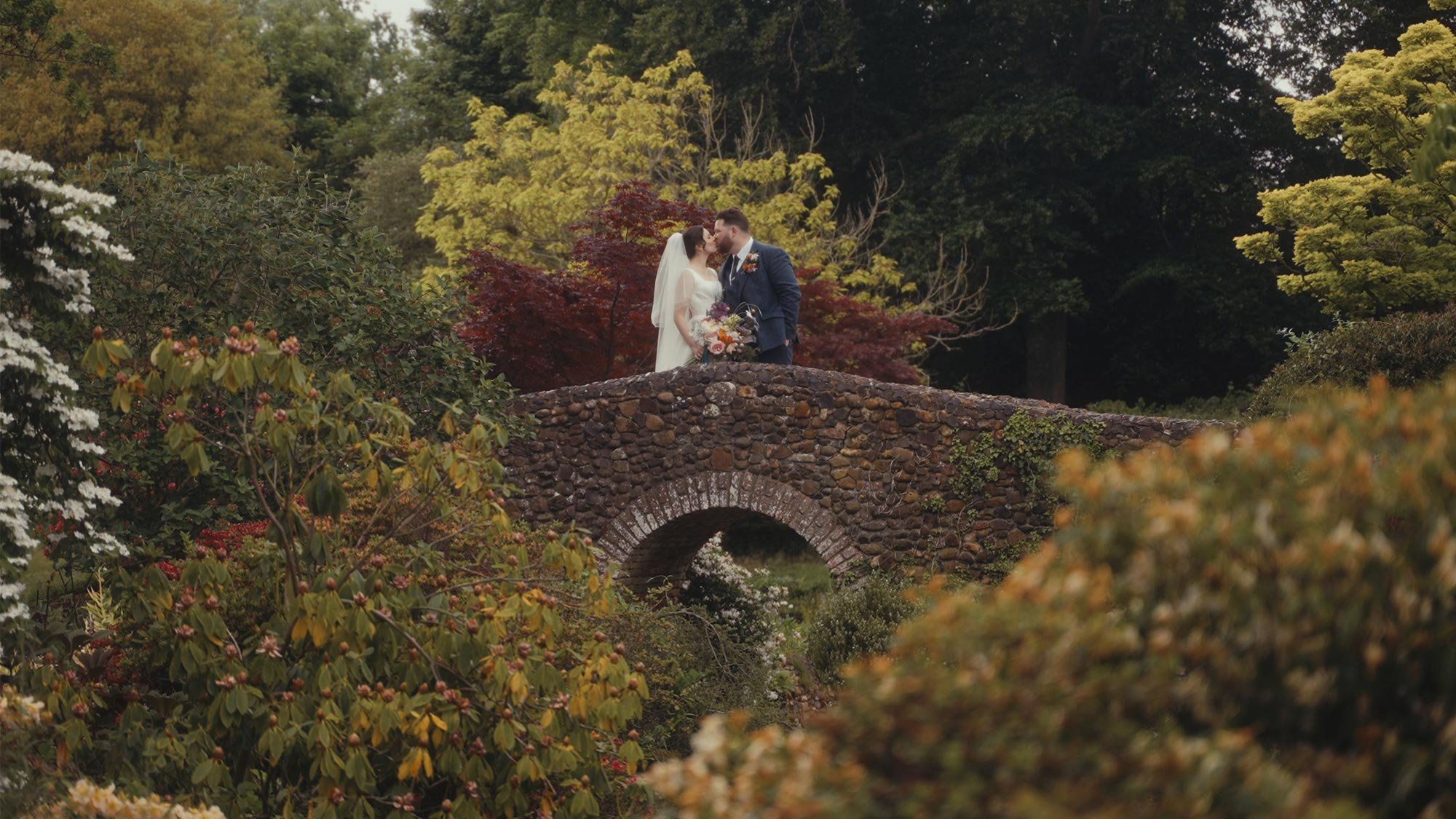 A bride and groom share a kiss on a stone bridge surrounded by lush, colorful garden foliage.