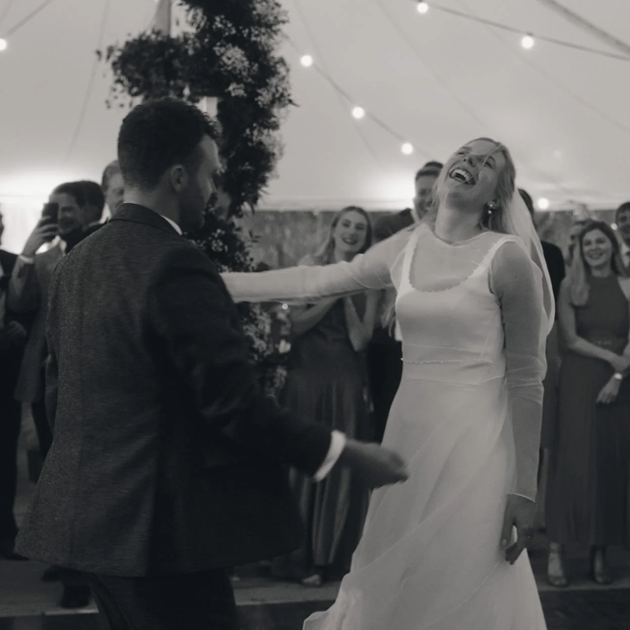 A bride and groom dancing joyfully under a tent with string lights, surrounded by smiling guests in formal attire.