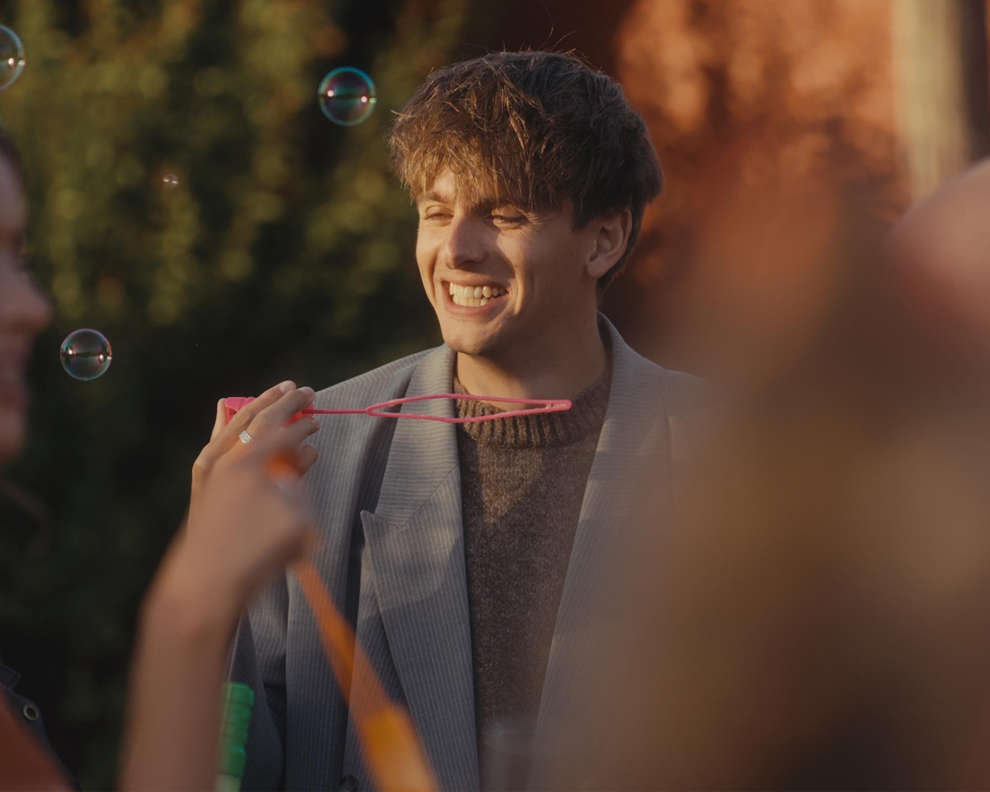 Charlie Pittman smiling outdoors during sunset, among people blowing bubbles, some holding bubble wands.