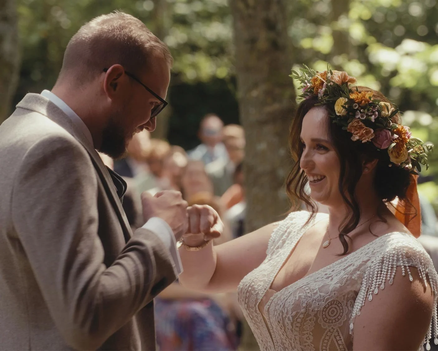 A couple fist-bumping during an outdoor wedding ceremony, with the bride wearing a floral crown and a lace dress.