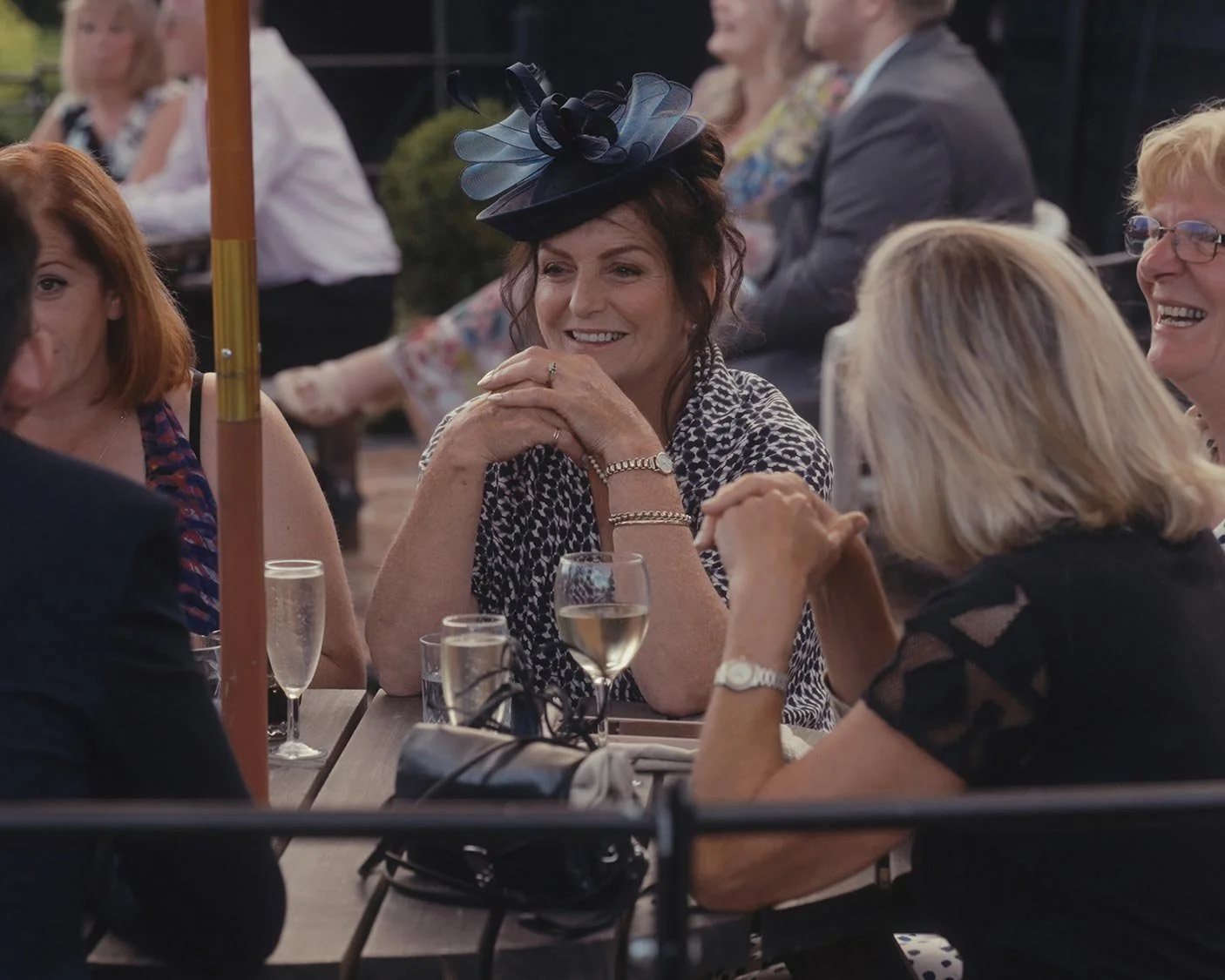 People gathered at an outdoor event, sitting around a table, engaged in conversation. One woman wears a fascinator, with drinks on the table.