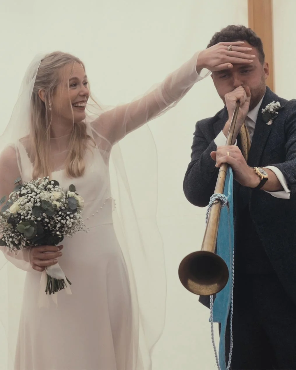 Bride laughing next to groom playing a long horn at wedding, bride holding bouquet.