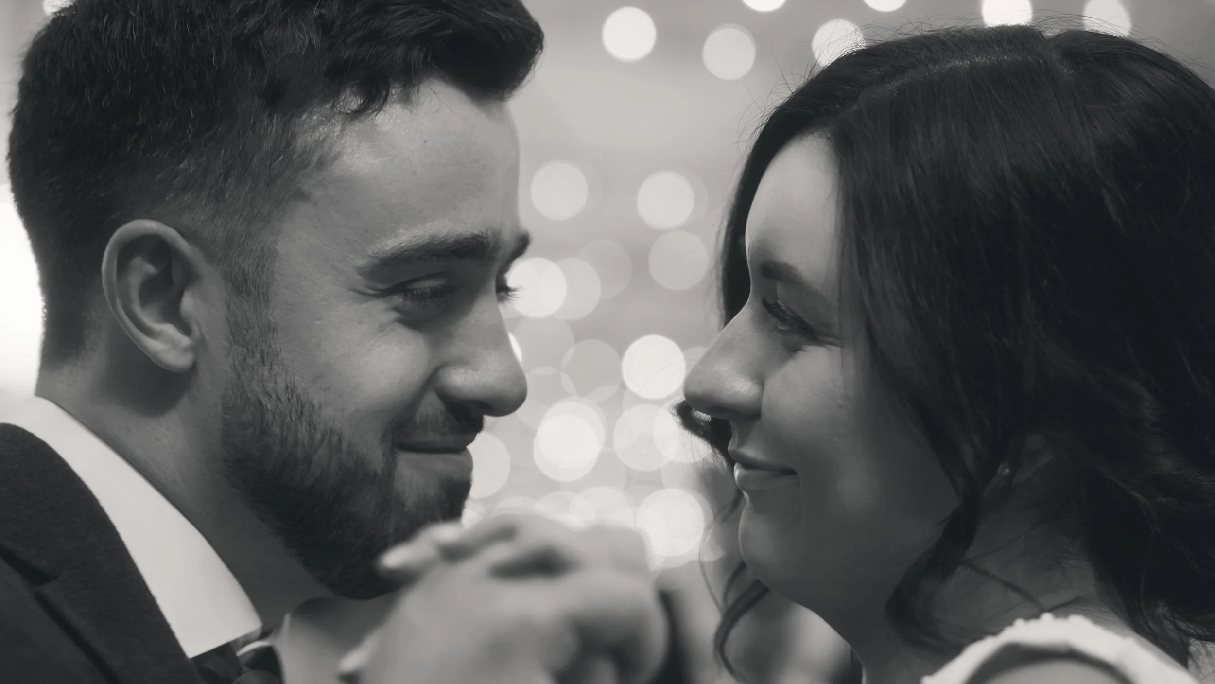 A black and white photo of a bride and groom close to each other, gazing into each other's eyes during their first dance, with blurred lights in the background.
