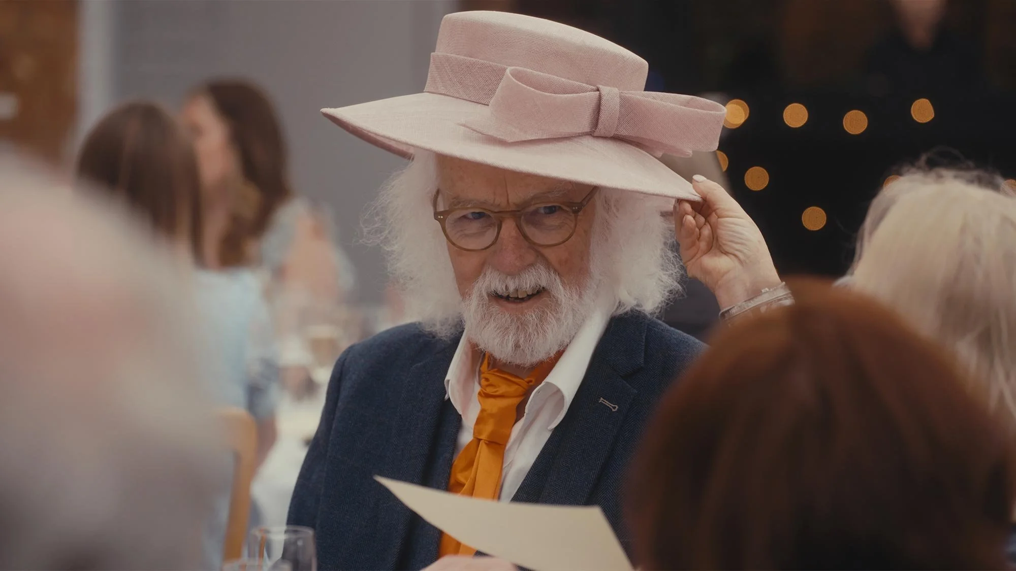 An elderly man having a pink ladies hat placed on his head at a wedding reception.