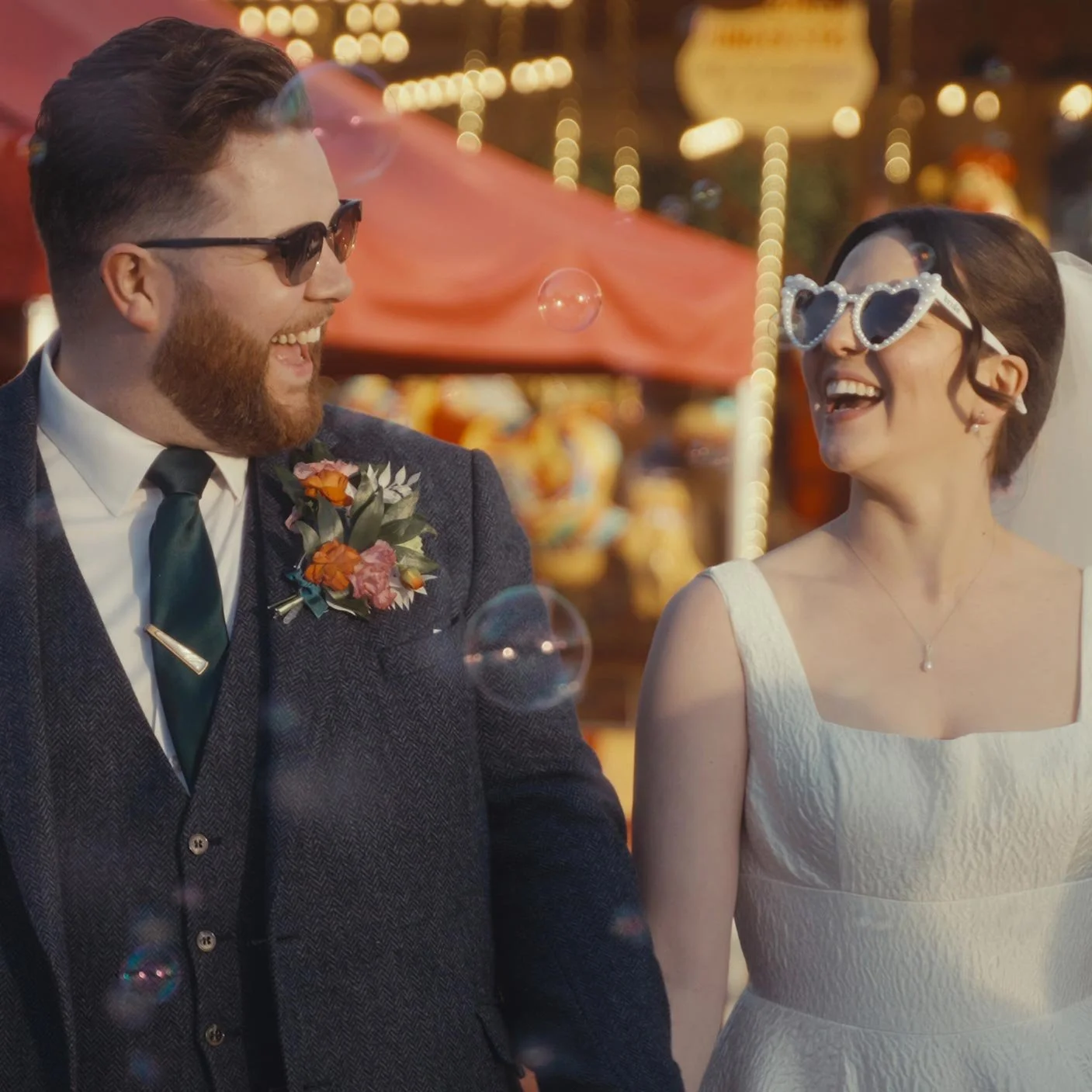 A smiling bride and groom walking together at a fairground, with the bride wearing playful heart-shaped sunglasses and a veil, and the groom in sunglasses and a suit with a boutonniere, as they enjoy their wedding day.