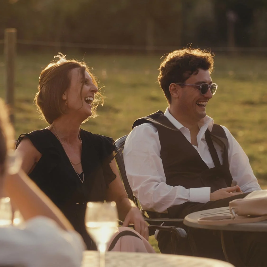 Group of people laughing outdoors at sunset, sitting at a table with drinks.