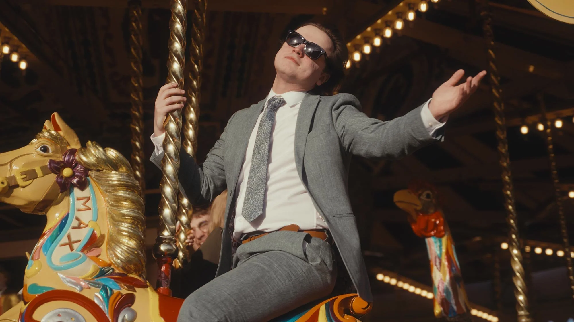 A man dressed in a gray suit, white shirt, and patterned tie, wearing sunglasses, is sitting on a colorful carousel horse with his arms raised and an expression of enjoyment.