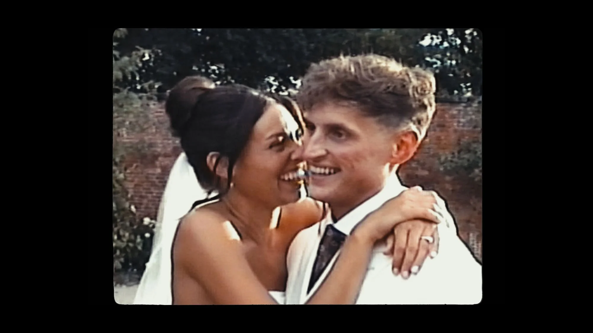 A young woman and a young man are smiling and embracing outdoors, with trees and a brick wall in the background.