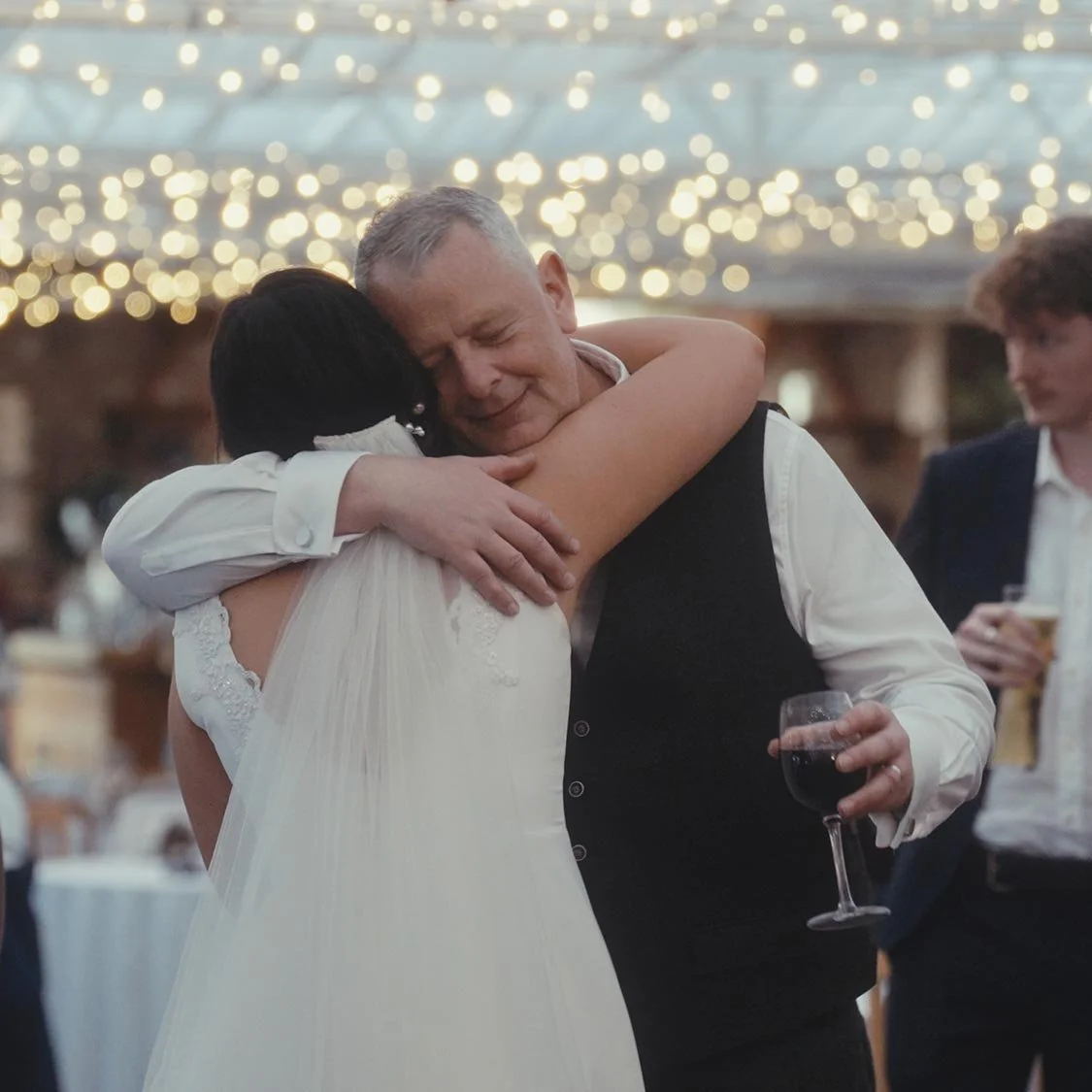 A daughter and her father hugging at a wedding, with people standing nearby holding drinks, under a ceiling decorated with string lights.