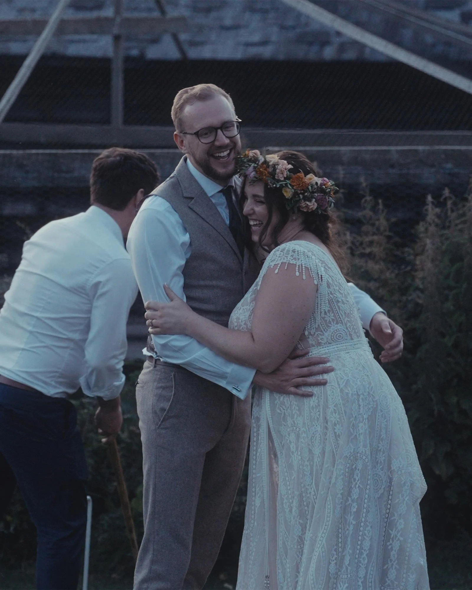 Bride and groom hugging outdoors, wearing wedding attire; man in background playing a game of croquet.