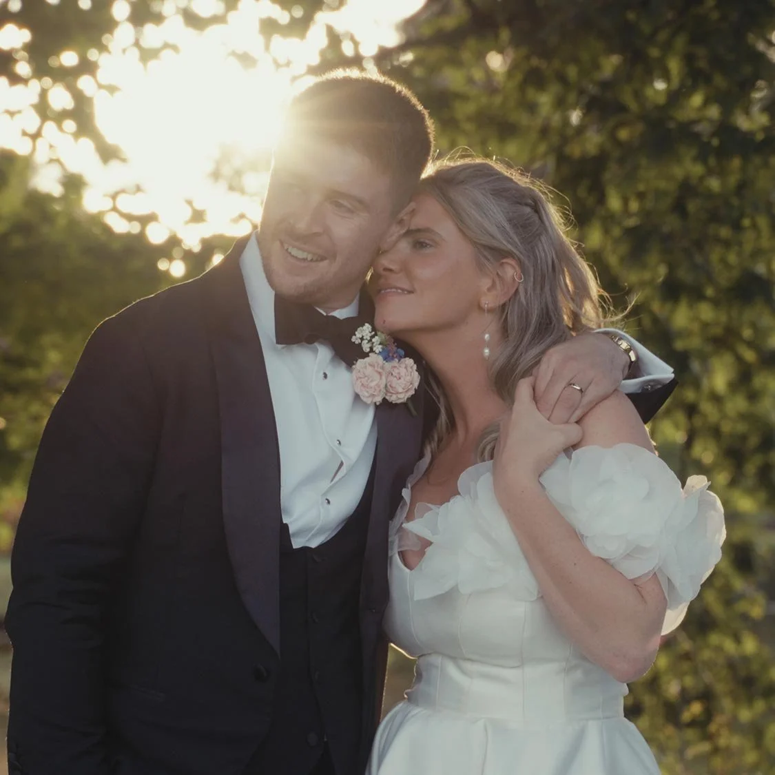 Bride and groom embracing outdoors with sunlight filtering through trees.
