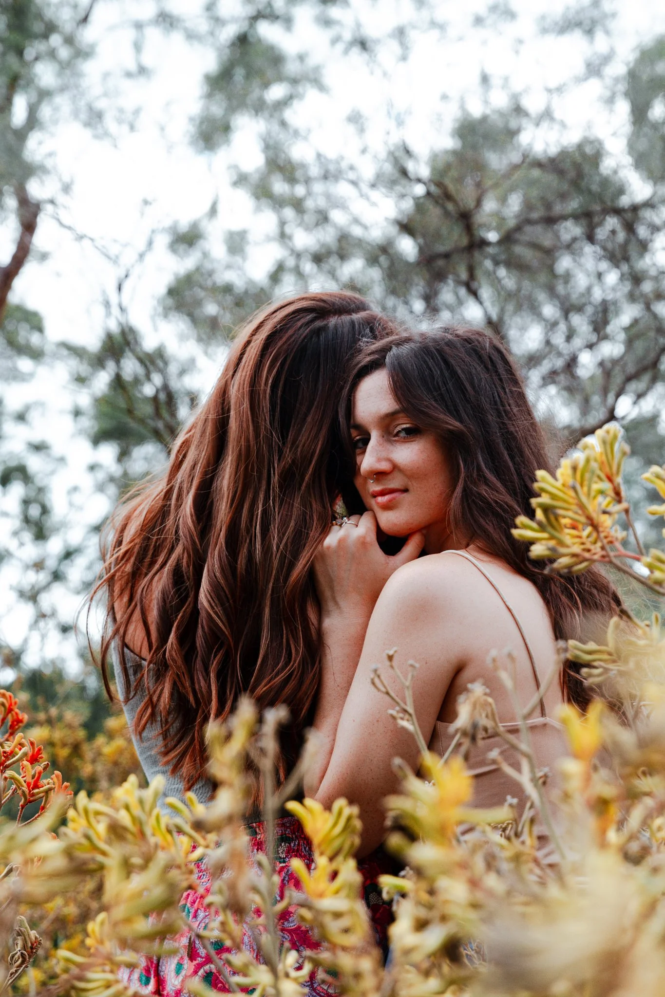 A female queer couple in a field of native kangaroo paw flowers, embracing with one back turned and one looking at the camera