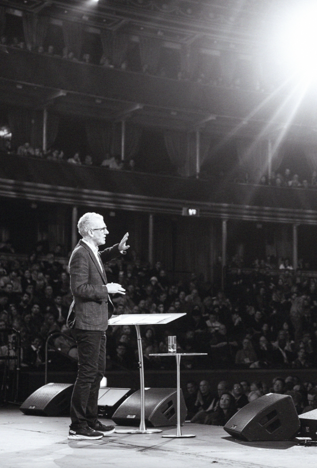 A man giving a speech or presentation on stage in front of an audience in a large theater or concert hall, with a music stand and a glass of water on a small table.