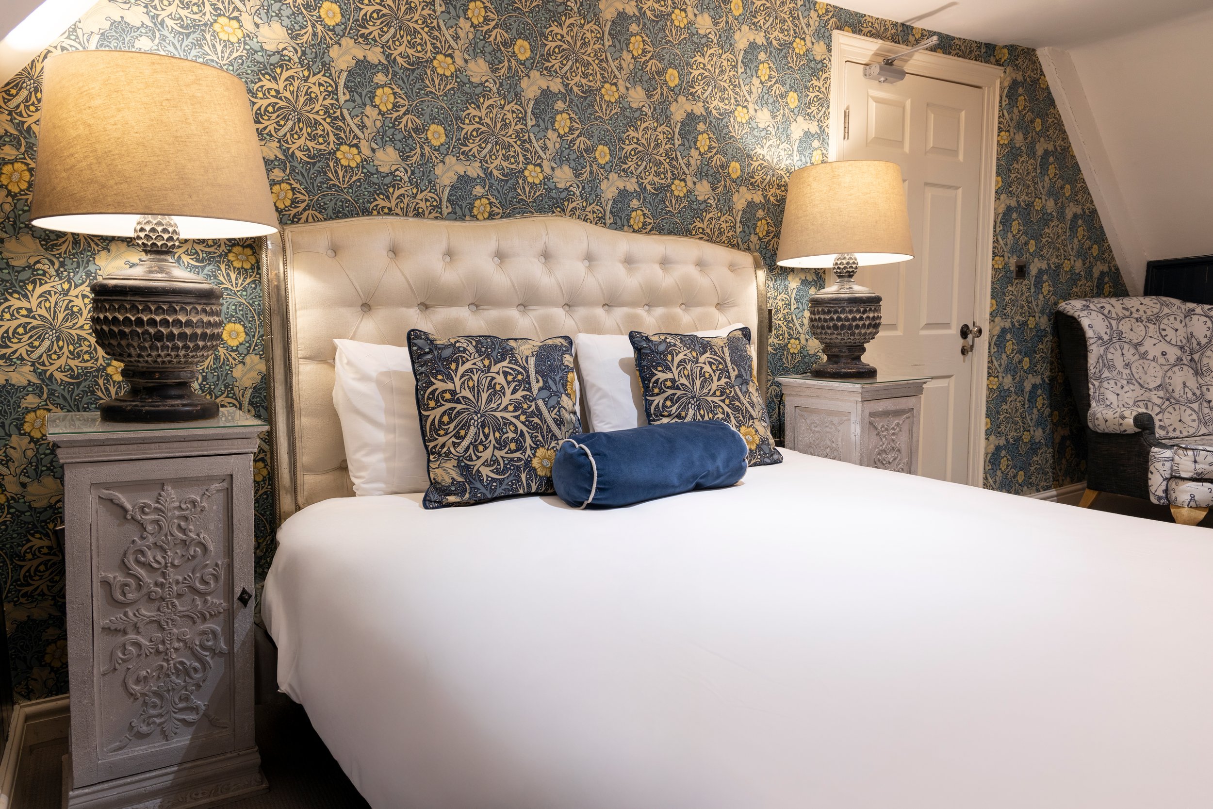 Elegant bedroom with a tufted headboard, decorative blue and gold pillows, floral wallpaper, wooden nightstands, and two lamps with beige lampshades.