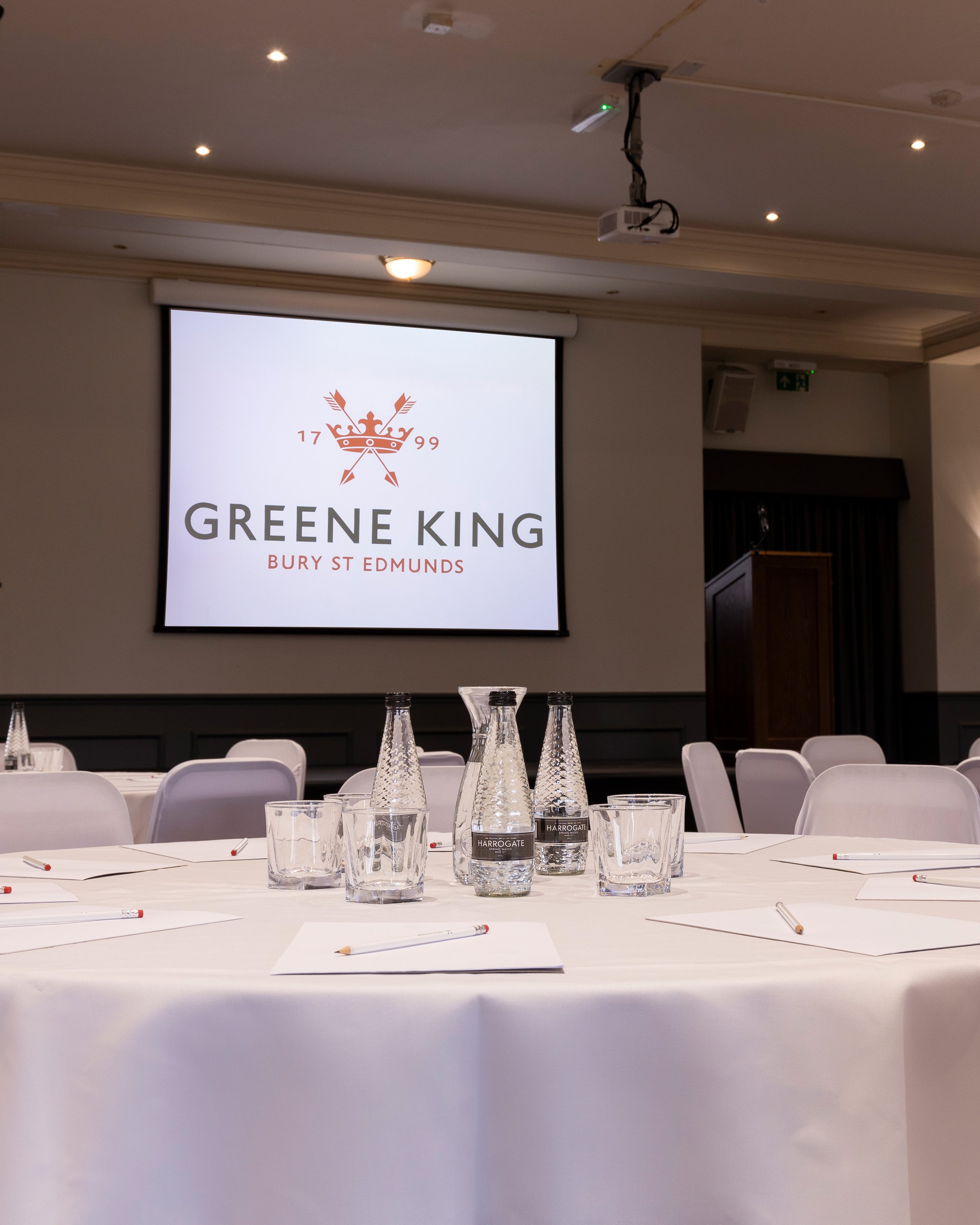 Conference setup with tables, chairs, glass bottles, notepads, and a projector screen displaying "Greene King Bury St Edmunds."